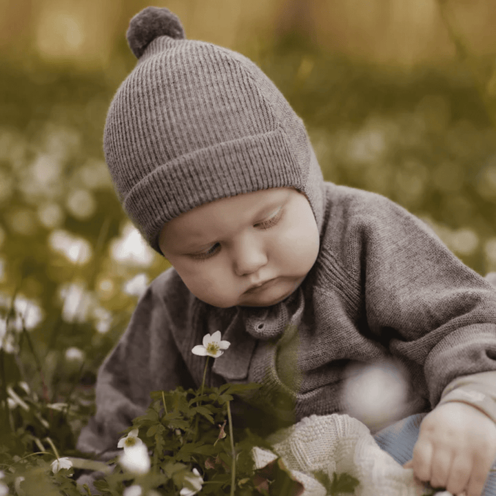 A Saga Copenhagen Merino Bonnet baby in a gray Saga Copenhagen Merino bonnet sitting in a field of flowers.