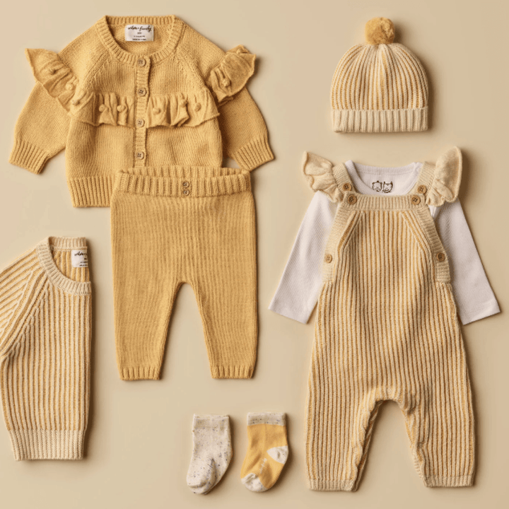 Collection of coordinated baby clothing and accessories laid out on a beige surface, including a yellow cardigan, striped pants, Wilson & Frenchy Knitted Ruffle Overalls for babies, a knitted hat, socks, and booties.