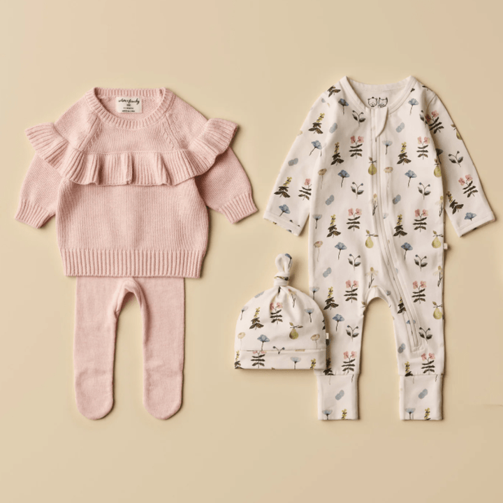 Two sets of Wilson & Frenchy Organic Baby Pyjamas displayed on a beige background, including a pink ruffled sweater with matching pants and a printed onesie with a coordinating hat featuring a hand-illustrated print.