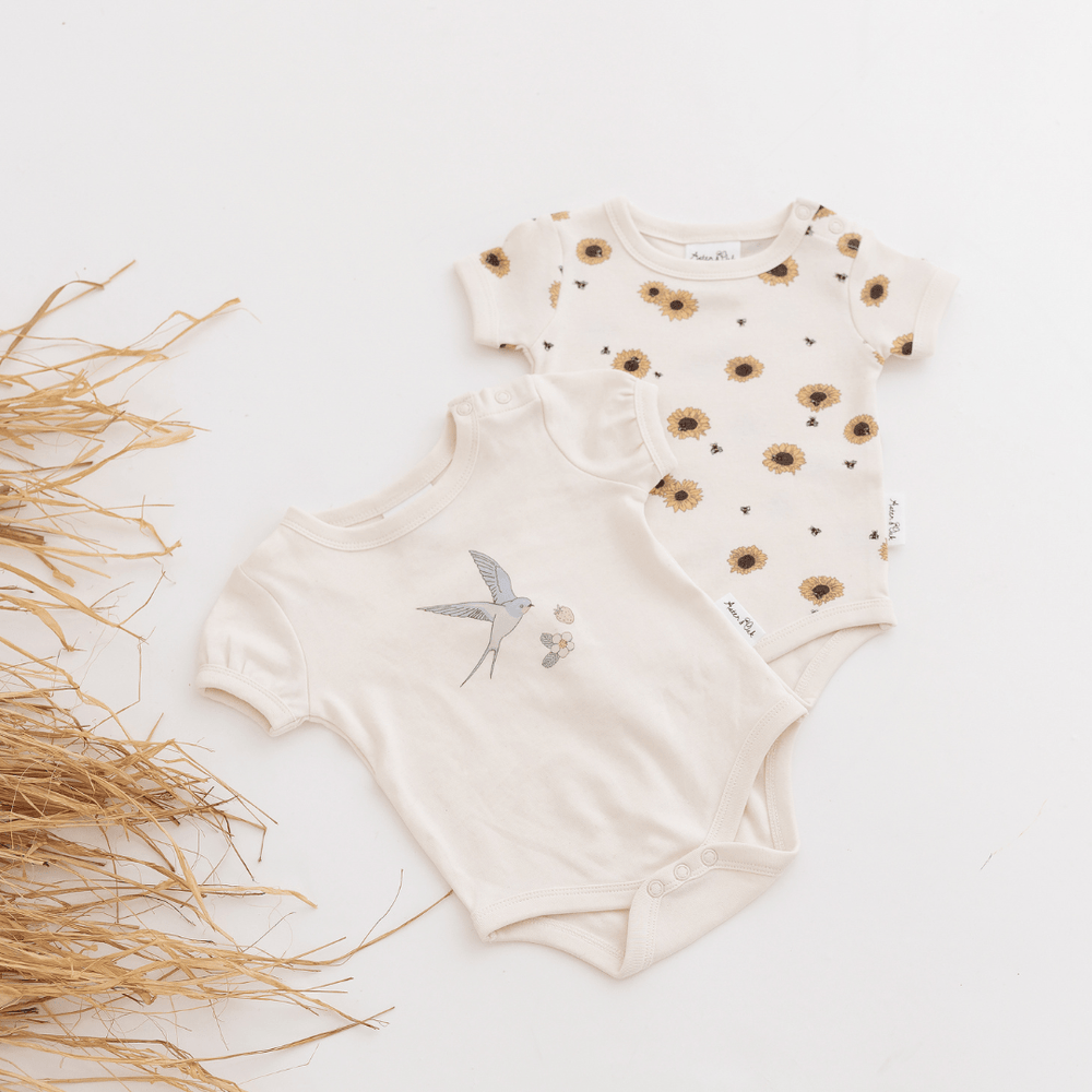 Two baby onesies lying on a white background with some dried grass beside them. One has puff sleeves and snaps at the crotch, featuring a delicate hand-illustrated swallow print on the chest, with a strawberry and strawberry flower, and the other has a sunflower print all-over.