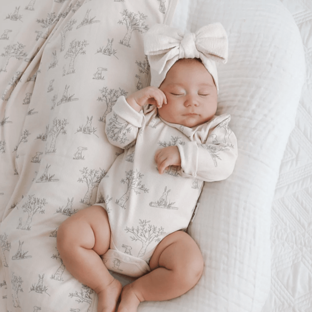 Newborn baby asleep on a white textured blanket, wearing an Aster & Oak Organic Bunny Luxe Rib Long-Sleeved Onesie with a bow headband.
