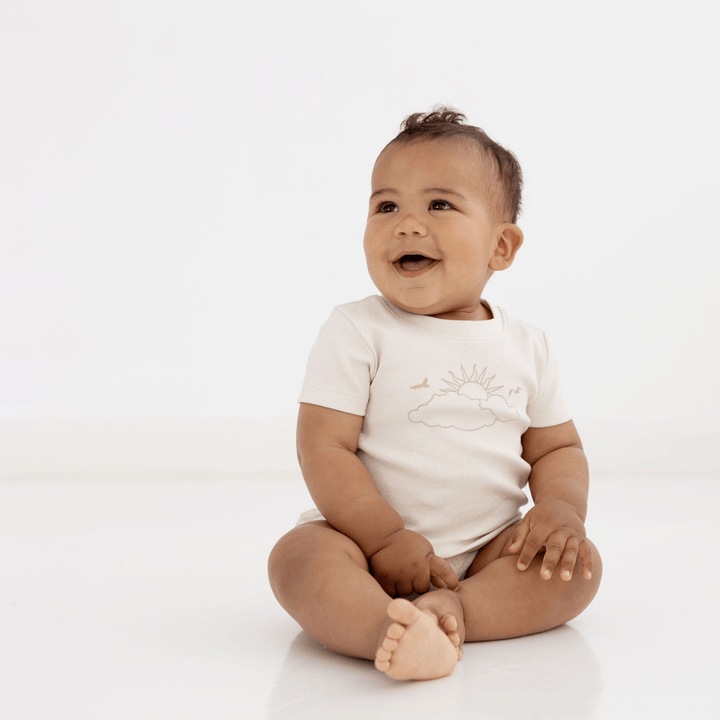 Adorable Baby With A Big Smile Sitting With Crossed Legs Wearing A Ribbed Cream T-Shirt Featuring the Aster & Oak Cloud Chaser Pattern (Sun & Cloud)
