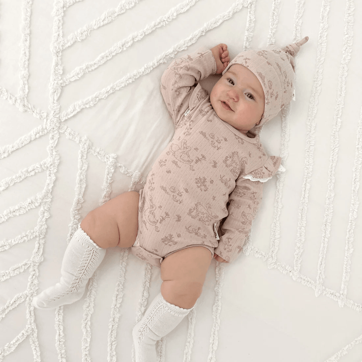 An infant wearing an Aster & Oak Organic Duck Family Flutter Onesie and a matching hat, lying on a white textured blanket.