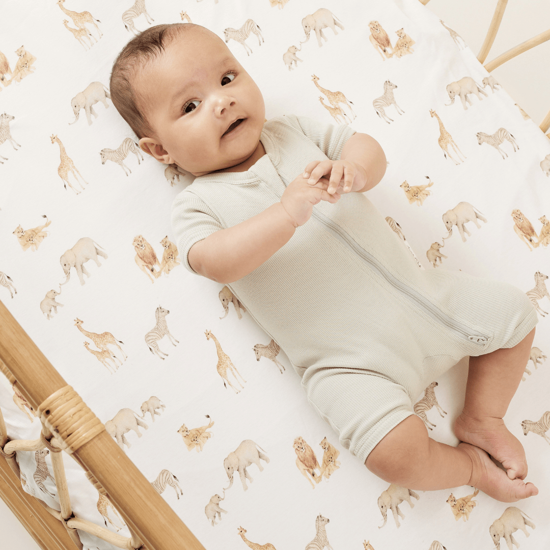 Smiling baby clasping his hands, lying in a rattan bassinet with a safari-themed sheet, wearing a sage-coloured baby zip romper