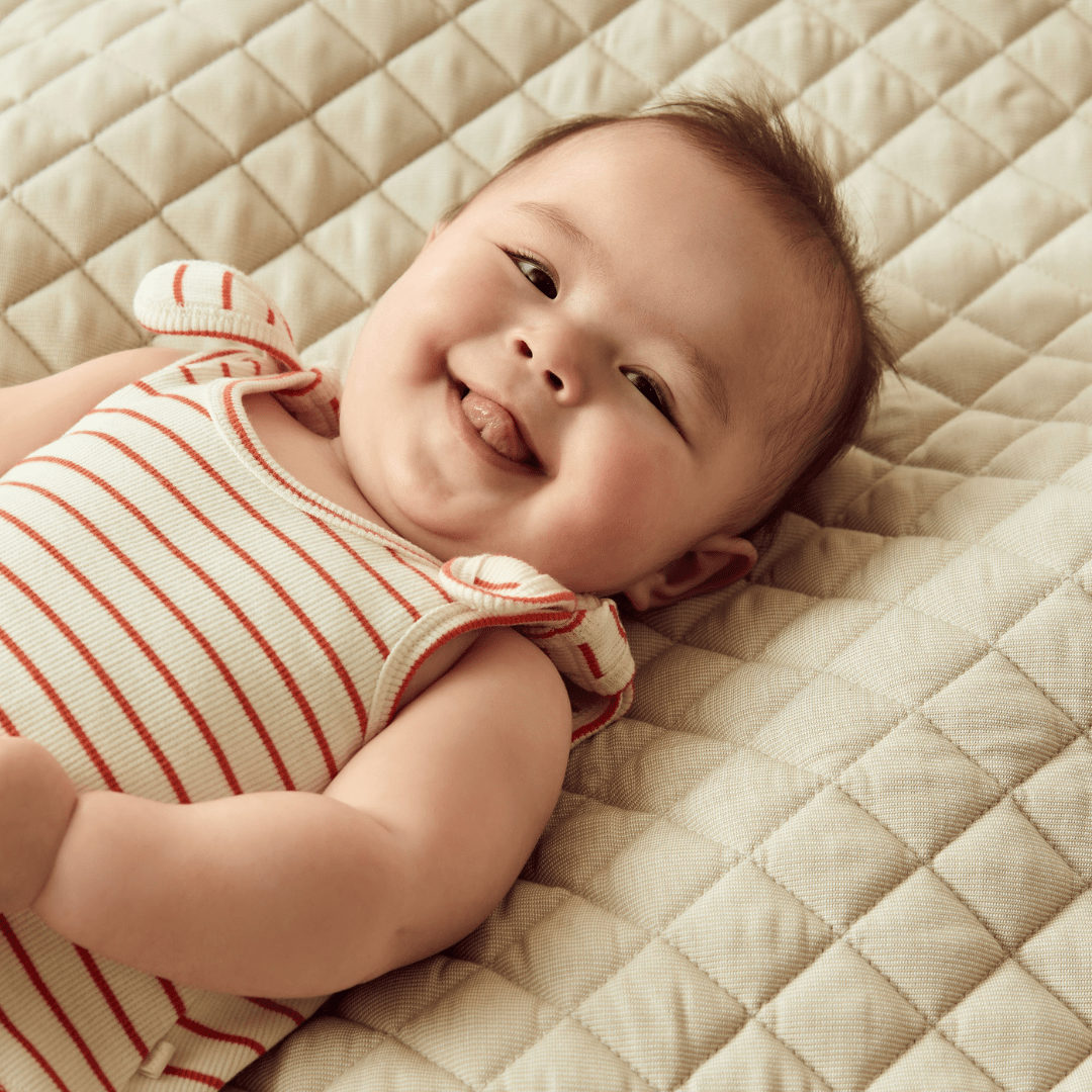 A Wilson & Frenchy baby smiles while laying on a bed, wearing a Wilson & Frenchy Organic Rib Stripe Tie Singlet made from GOTS-certified organic cotton.