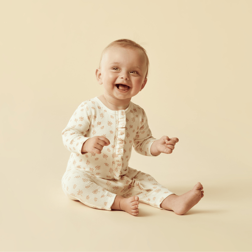 A smiling baby sitting down in a Wilson & Frenchy Organic Waffle Ruffle Zipsuit against a neutral background.