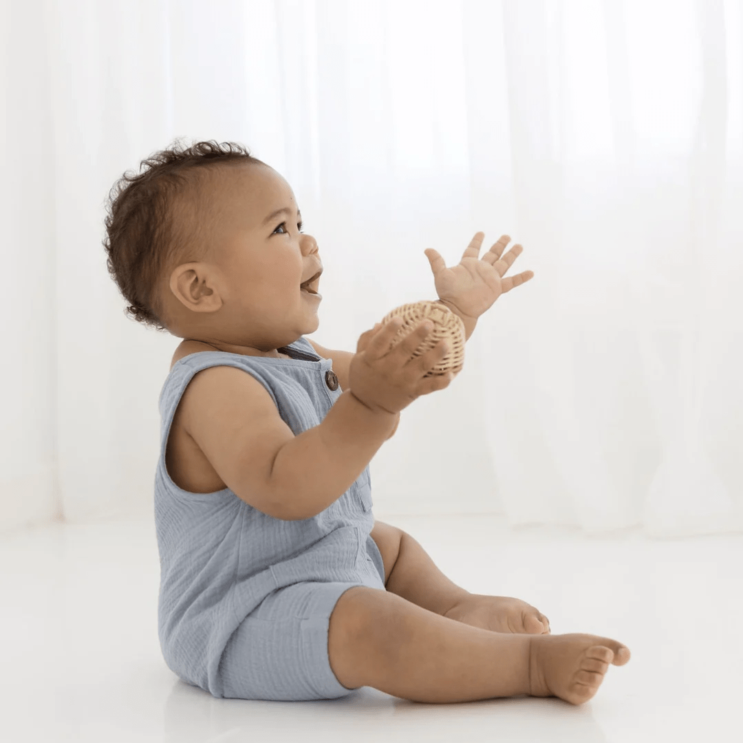 A baby is sitting on the floor and reaching for the Aster & Oak Organic Slate Blue Muslin Romper - LUCKY LAST - 6-12 MONTHS ONLY during the summer.