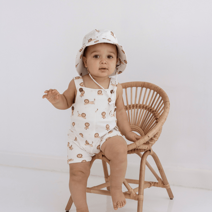 An adorable baby sitting on a wicker chair wearing a hat and Aster & Oak Organic Cotton Lion Overalls, made of organic cotton.