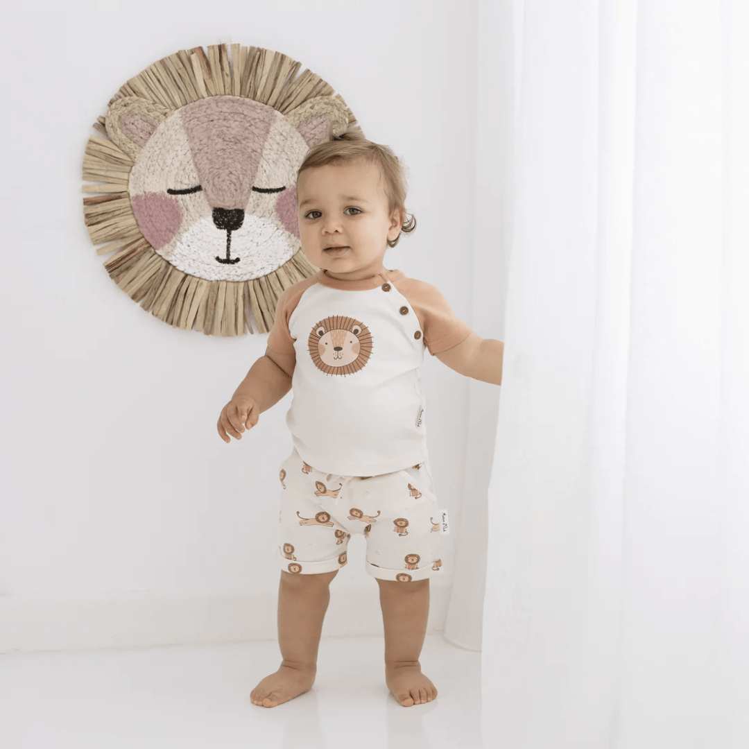 A baby wearing an Aster & Oak certified organic cotton Lion Print Top stands in front of a curtain with a lion on it.