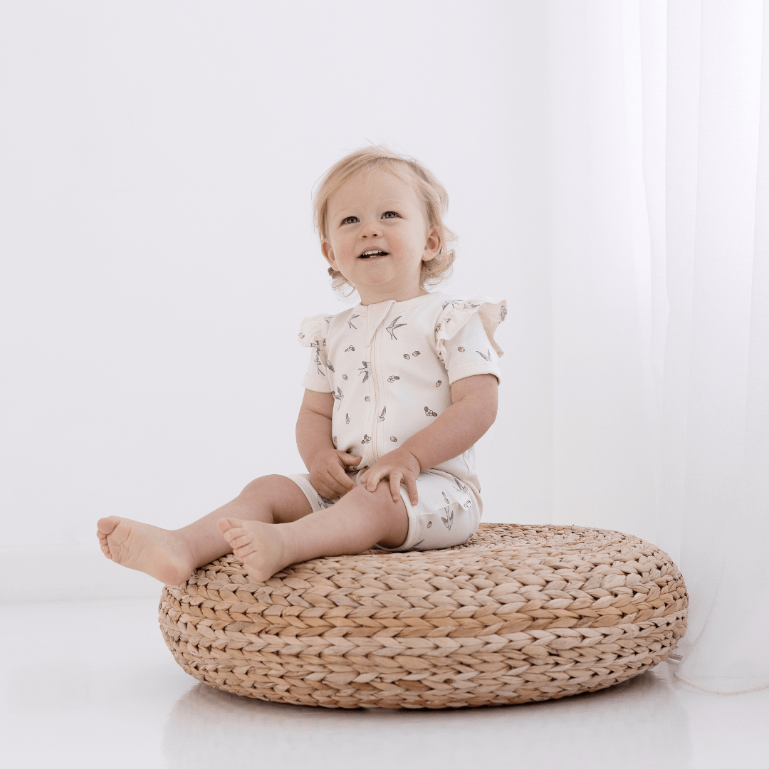 A baby sitting on a wicker basket wearing an Aster & Oak Organic Cotton Flutter Sleeve Zip Romper in front of a white wall.