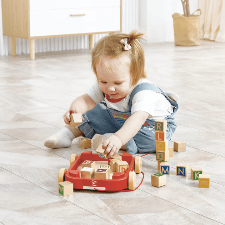 Happy-Girl-Stacking-Blocks-Into-Hape-Pull-Along-Cart-With-Stacking-Blocks-Naked-Baby-Eco-Boutique
