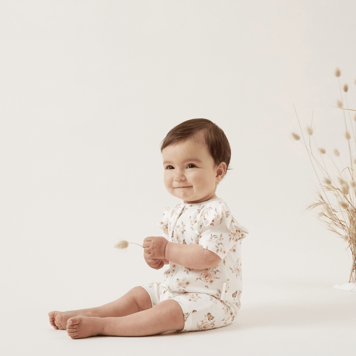A baby sitting on the floor in a delicate white Aster & Oak Organic Cotton Flutter Sleeve Zip Romper.
