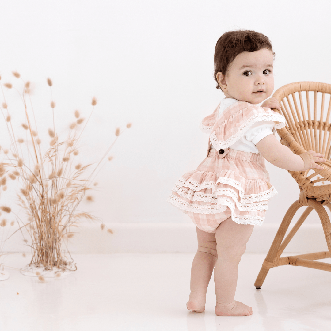 Beautiful baby with dark hair wearing pink and white ruffle gingham playsuit, standing holding on to a a rattan chair, with dried flowers in the background