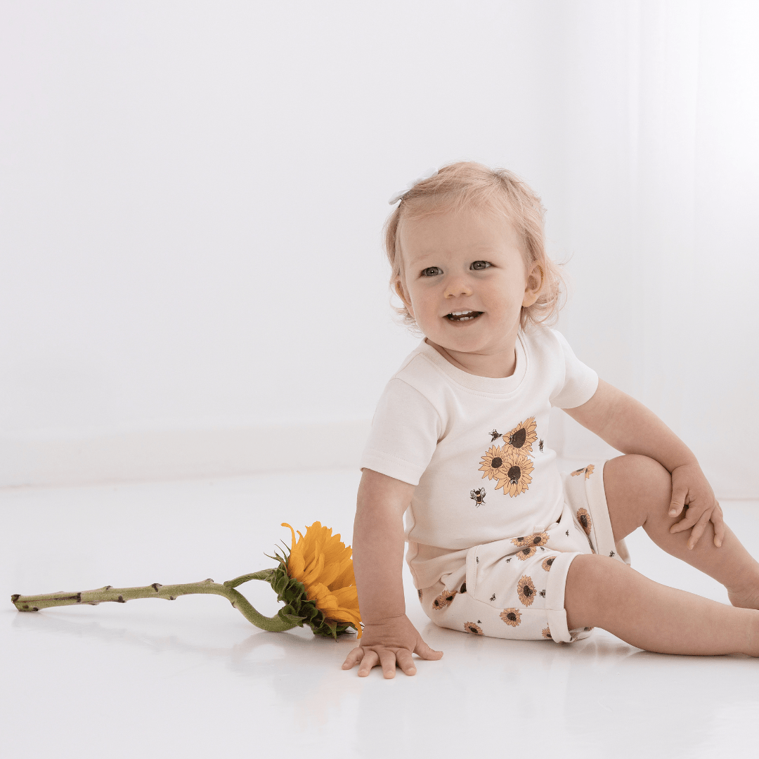 An adorable baby, wearing Aster & Oak organic cotton harem shorts with an adjustable drawstring waist tie, happily sits on the floor next to a sunflower.