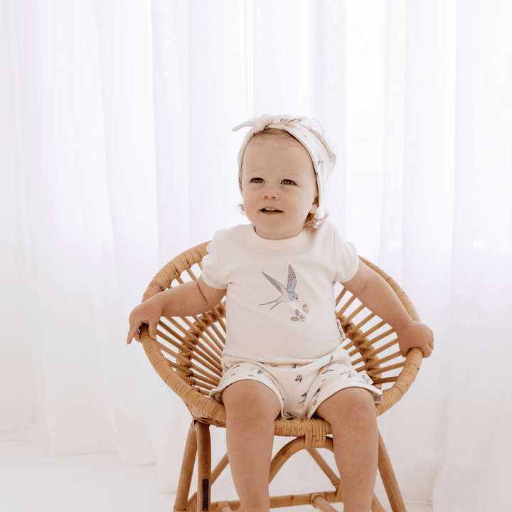 Toddler girl sitting in a rattan chair wearing a watercolour swallow-print headband with a bow on it, and matching shorts and t-shirt