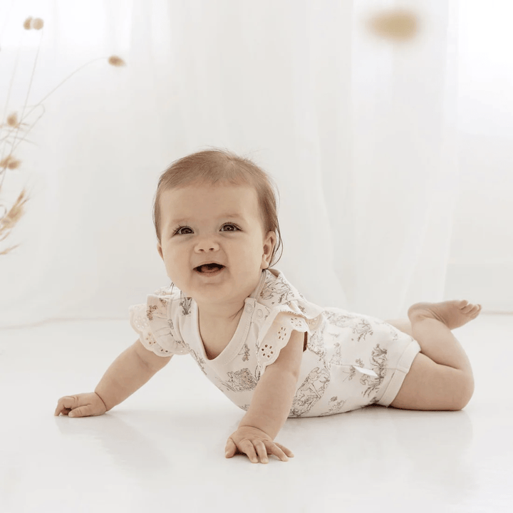 A baby is laying on the floor in a white Aster & Oak Organic Cotton Meadow Flutter Onesie with flutter sleeves.