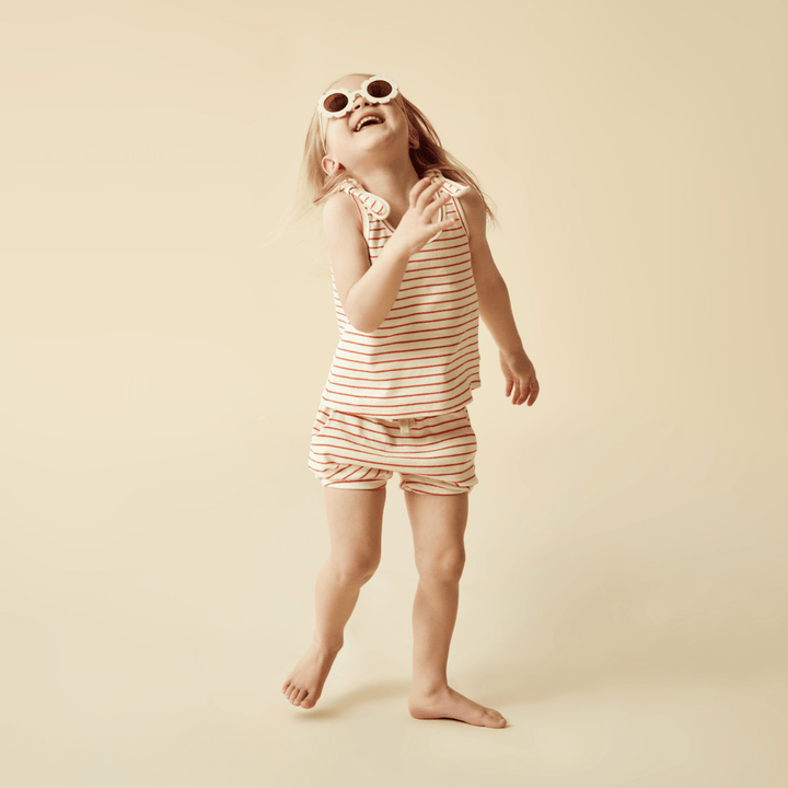 A little girl wearing sunglasses and a striped romper completes her outfit with the Wilson & Frenchy Organic Rib Stripe Tie Kids Singlet (Multiple Variants) from Wilson & Frenchy. Made with GOTS-certified organic cotton, this singlet adds the perfect finishing touch to her look.