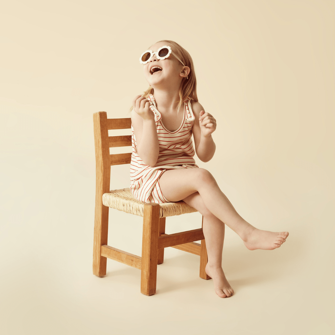 A little girl wearing sunglasses, sitting on a wooden chair, completes her summer outfit with the Wilson & Frenchy GOTS-certified organic cotton Wilson & Frenchy Organic Rib Stripe Tie Kids Singlet (Multiple Variants) and the Summer Stripe.