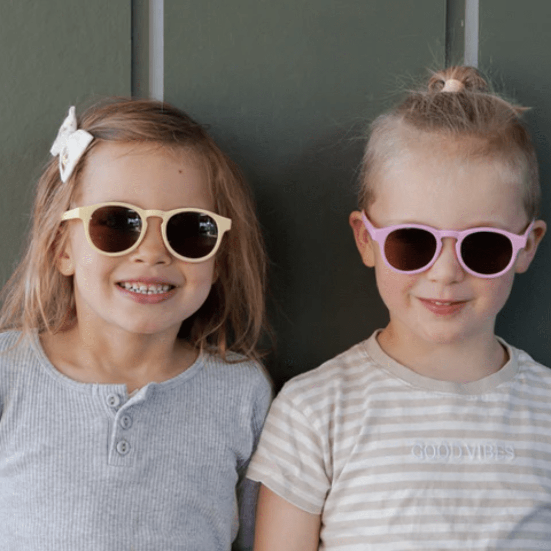 Two little girls wearing Zazi Shades - Kids 3+ Years sunglasses in front of a green wall on sunny summer days.