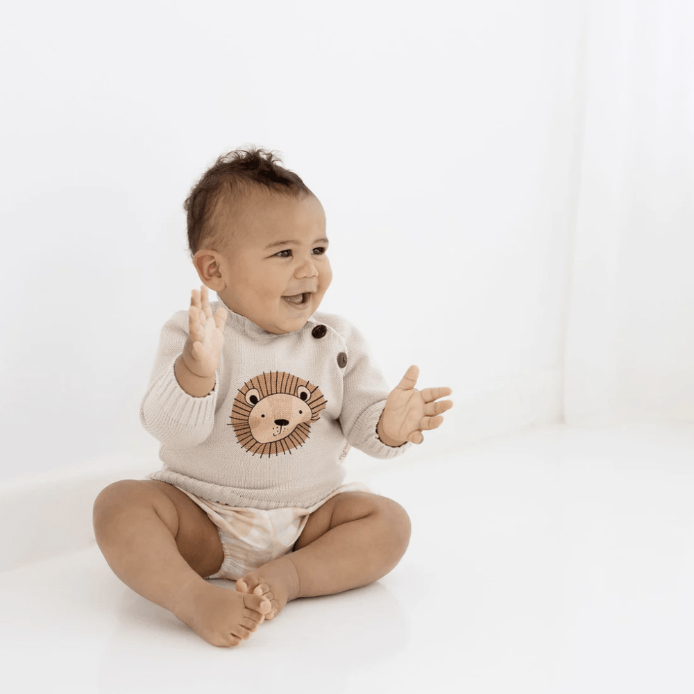 A baby is sitting on the floor in a cozy Aster & Oak Organic Lion Knit Jumper, made of organic cotton, with his hands up.
