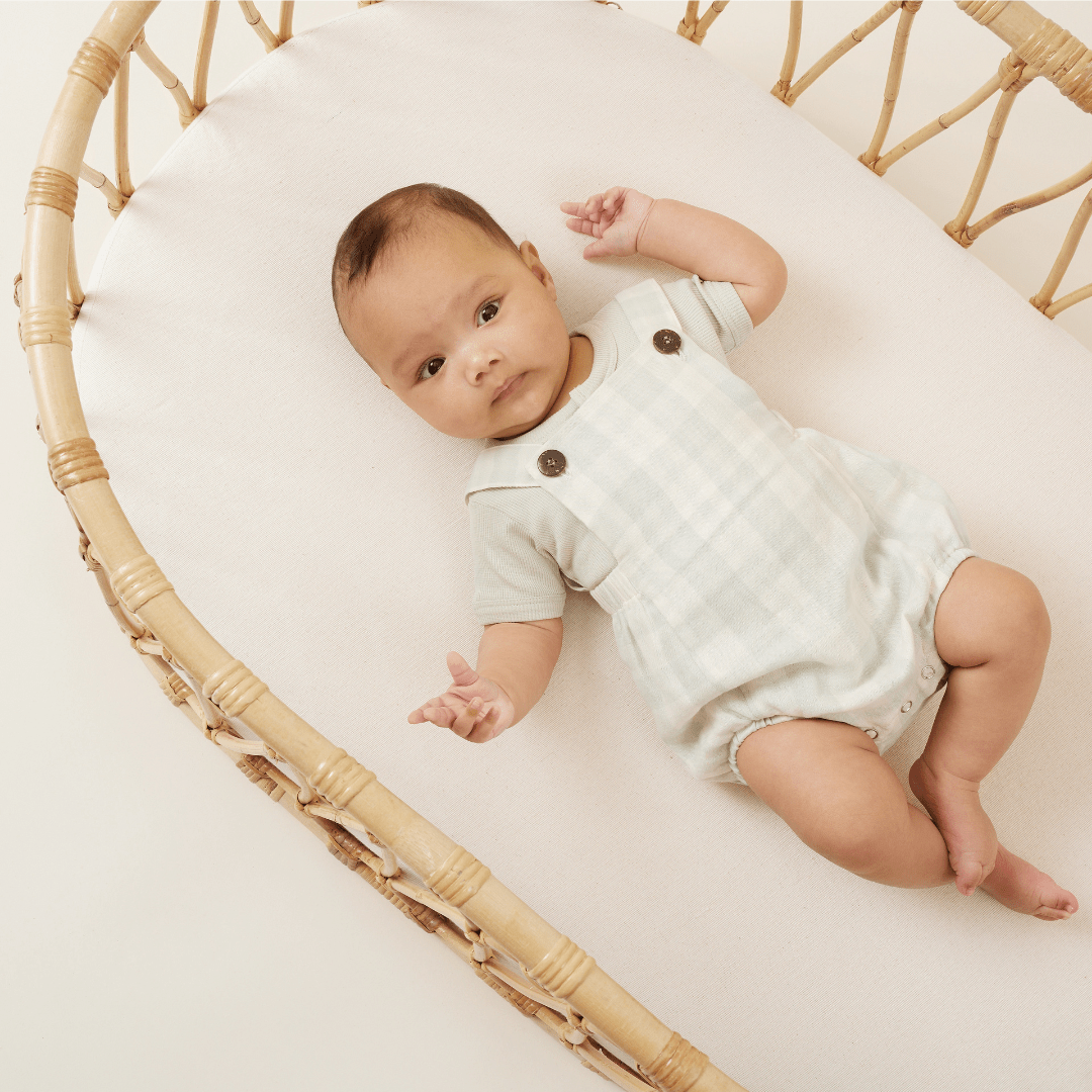 Beautiful baby boy lying in rattan bassinet wearing a sage-coloured onesie underneath the sage gingham playsuit with coconut buttons