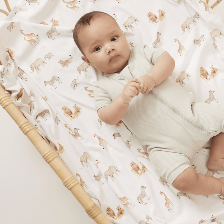 Baby boy lying on top of a safari-themed blanket, in a rattan cot, wearing a sage green ribbed baby zipsuit, holding on to his hands