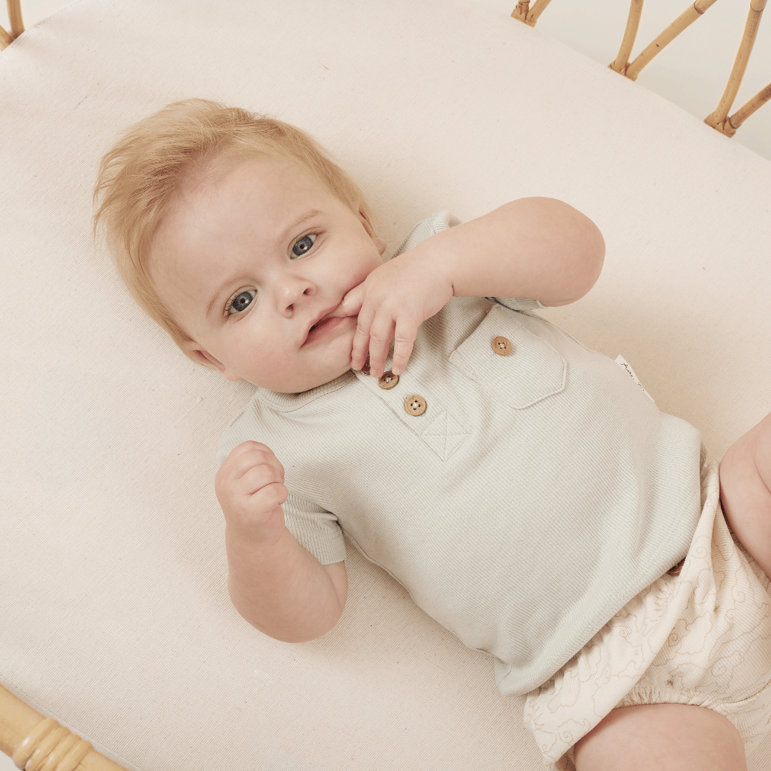 A baby lying on a wicker surface, dressed in an Aster & Oak Organic Rib Henley Onesie, with one hand near the mouth.