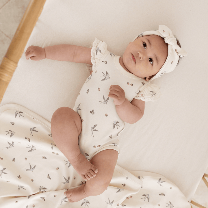 A baby laying on a bed wrapped in an Aster & Oak Organic Cotton Baby Swaddle Wrap with a hand-illustrated print and wearing a headband.