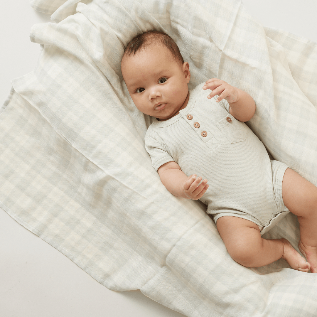 A baby wearing an Aster & Oak Organic Rib Henley Onesie lying on an organic cotton checked blanket, gazing upward.