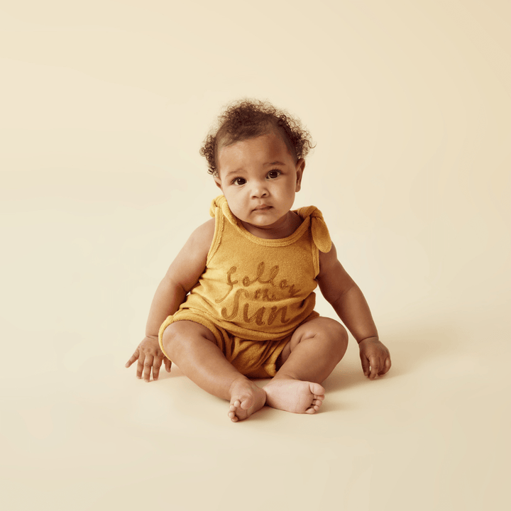 A baby in a Wilson & Frenchy Organic Terry Tie Singlet (Multiple Variants), made from GOTS-certified organic fabric, sitting on a beige background.