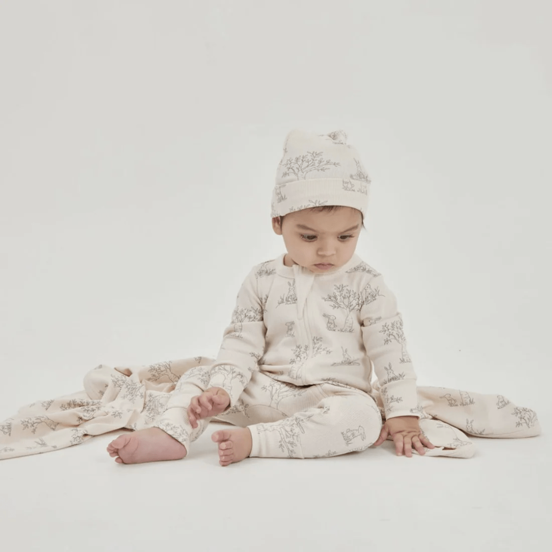 A newborn dressed in a matching Aster & Oak Organic Bunny Luxe Rib Long-Sleeved Zip Romper and hat with a print design, sitting on a white background.