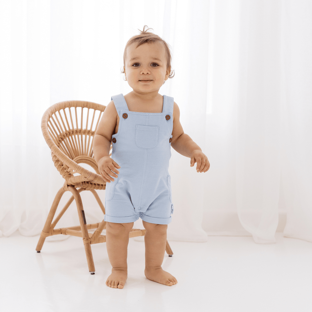 Toddler standing in a bright room wearing blue, gender-neutral Aster & Oak Organic Cotton Chambray Overalls next to a wooden chair.