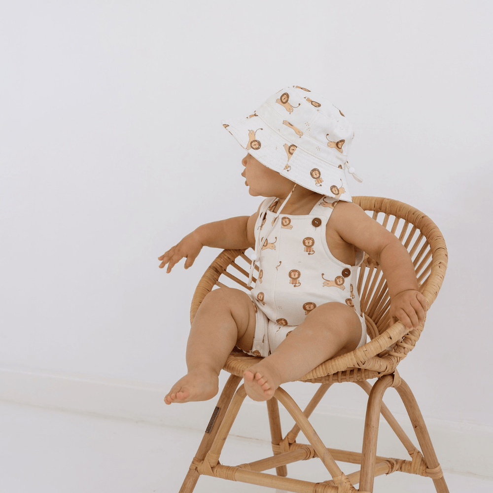 A baby sitting in a wicker chair wearing a hat, Aster & Oak Organic Cotton Lion Overalls - LUCKY LAST - 0-3 MONTHS ONLY on final sale.
