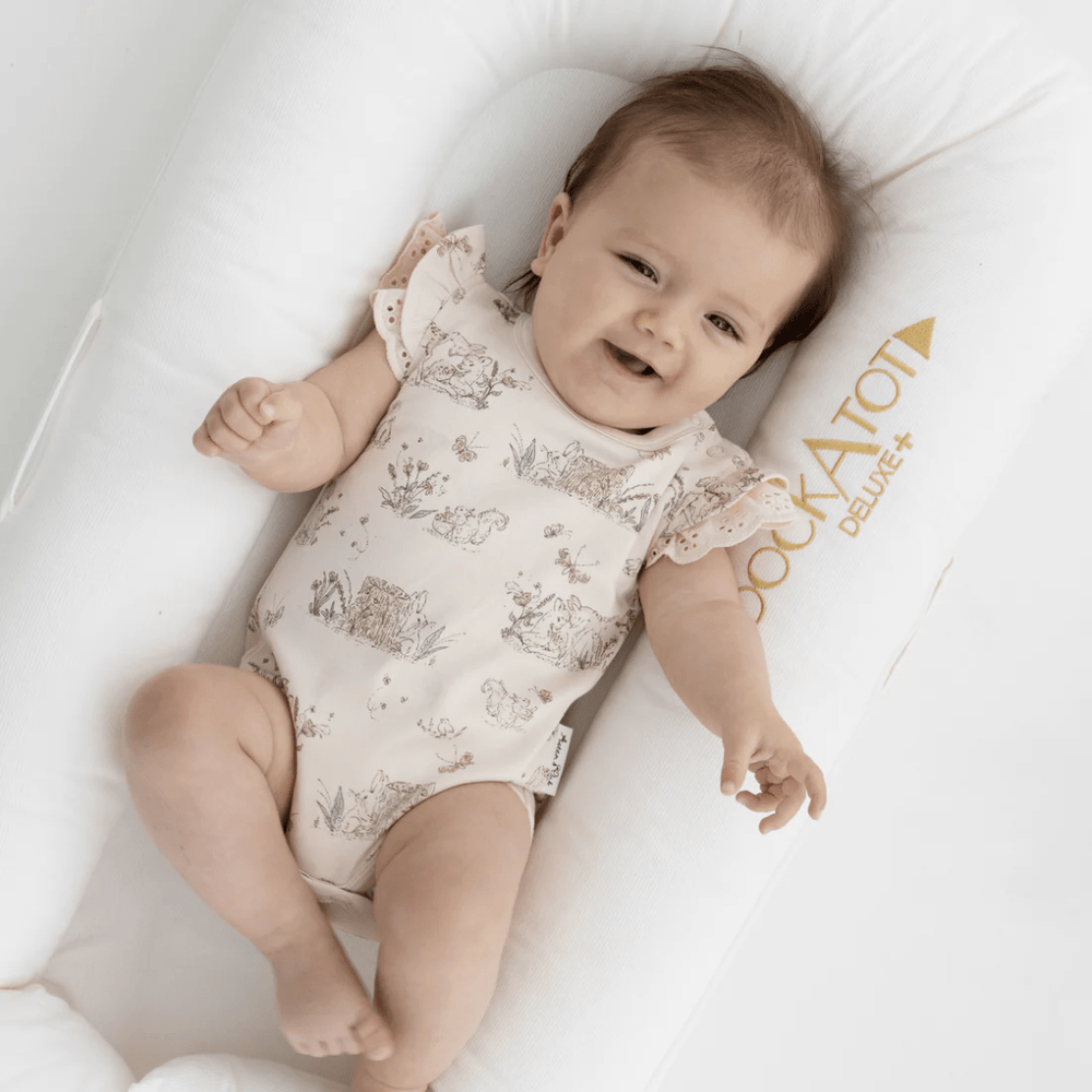 An adorable baby dressed in an Aster & Oak Organic Cotton Meadow Flutter Onesie - LUCKY LASTS - 1 YEAR ONLY, resting peacefully on a white pillow in a baby carrier.