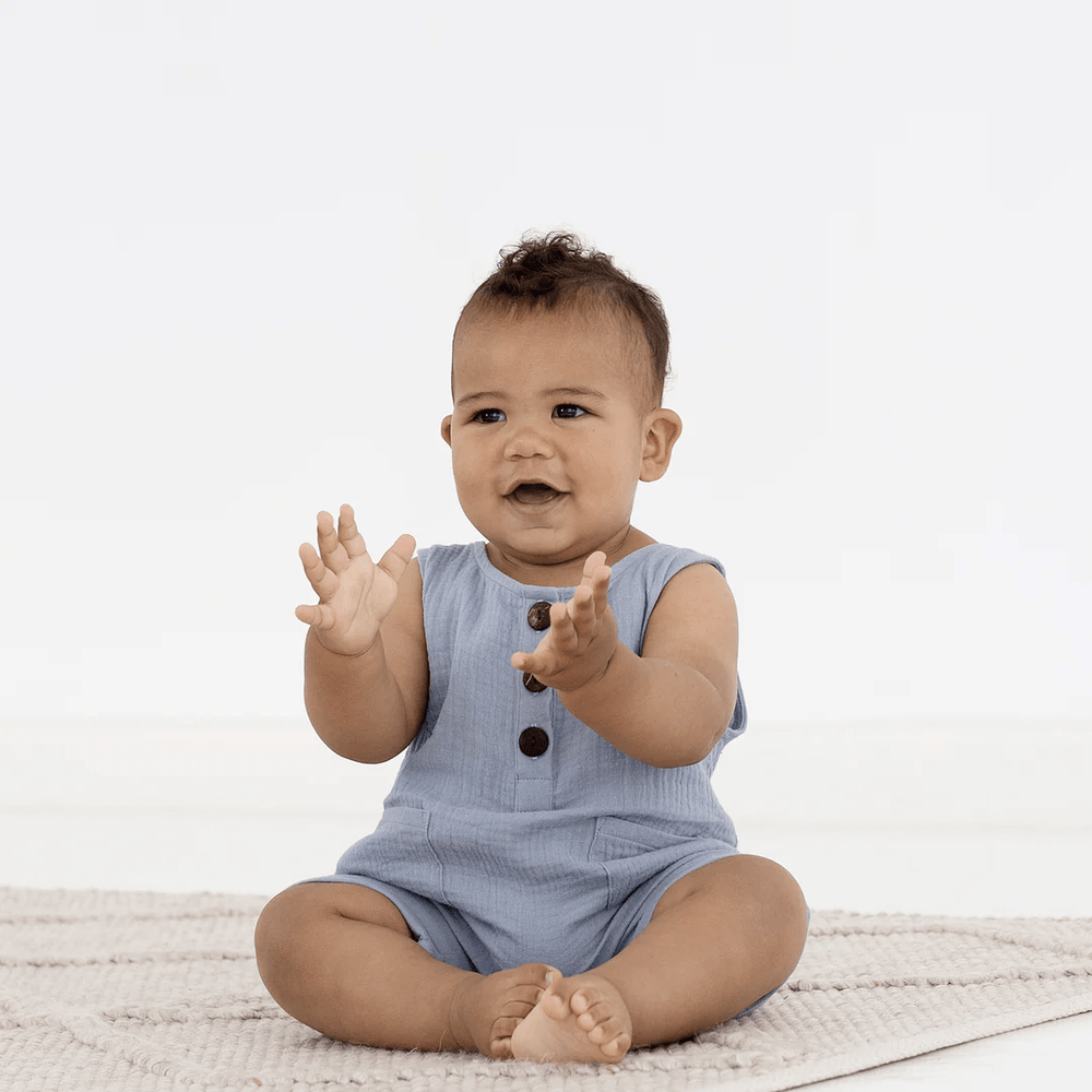 A baby sitting on an Aster & Oak Organic Slate Blue Muslin Romper with his hands up.