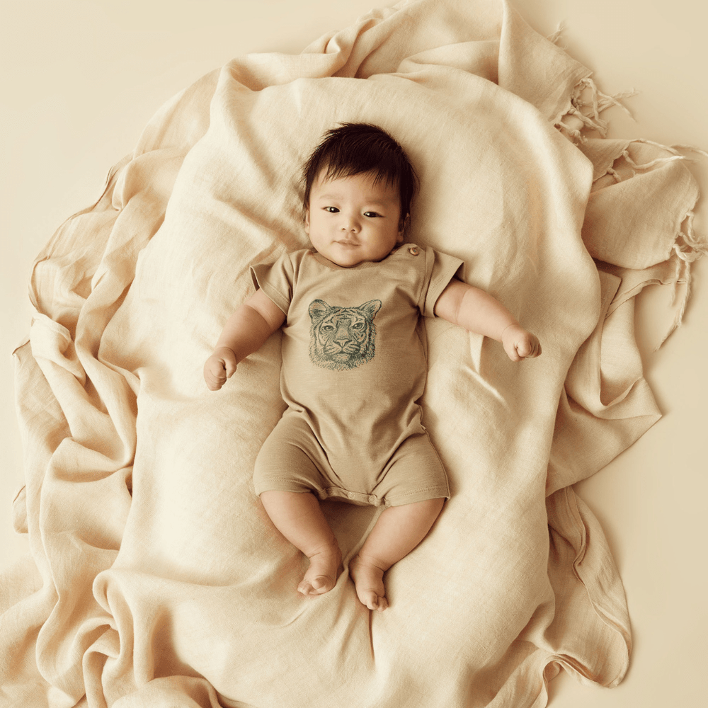 A baby wearing the Wilson & Frenchy Organic Leo the Lion Boyleg Growsuit lays on a blanket with a tiger on it.