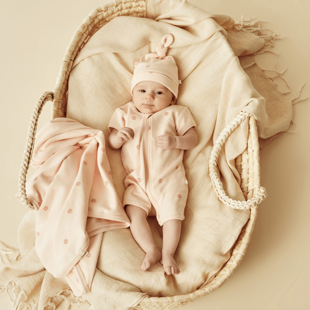 A baby is laying in a basket with a blanket and a Wilson & Frenchy Organic Rib Knot Hat.