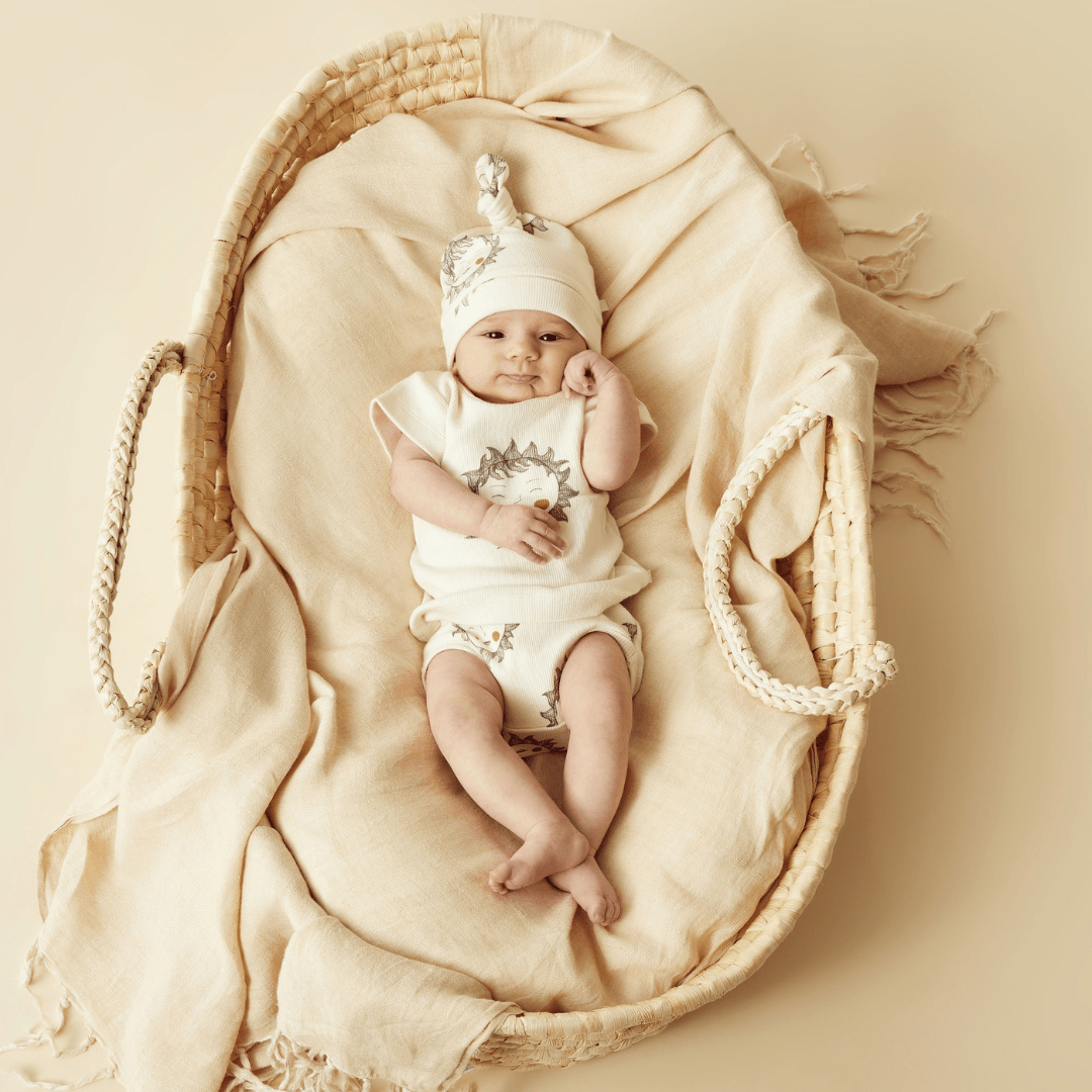 A baby nestled in a basket on a beige background, wearing the Wilson & Frenchy Organic Rib Knot Hat by Wilson & Frenchy.
