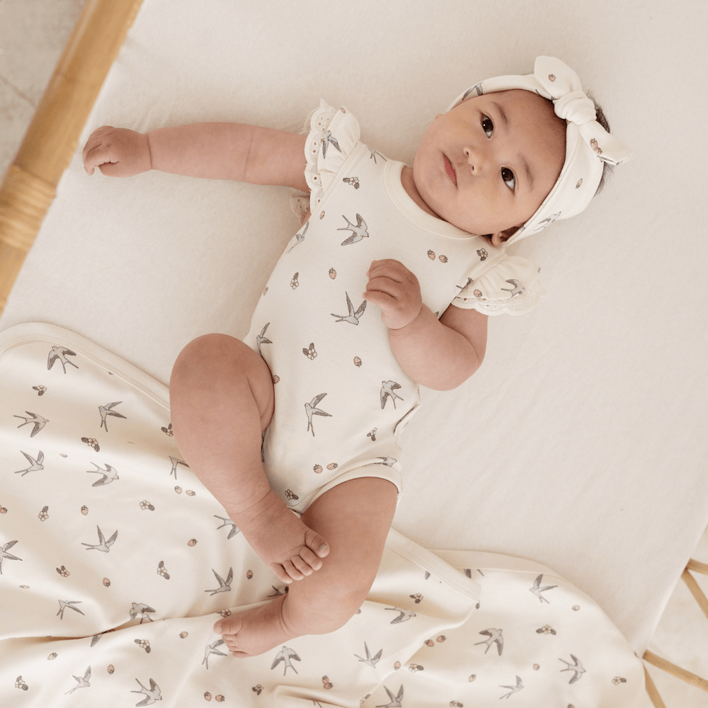 Baby girl lying in a rattan cot wearing a onesie featuring a delicate swallow and strawberry print, with matching headband and blanket