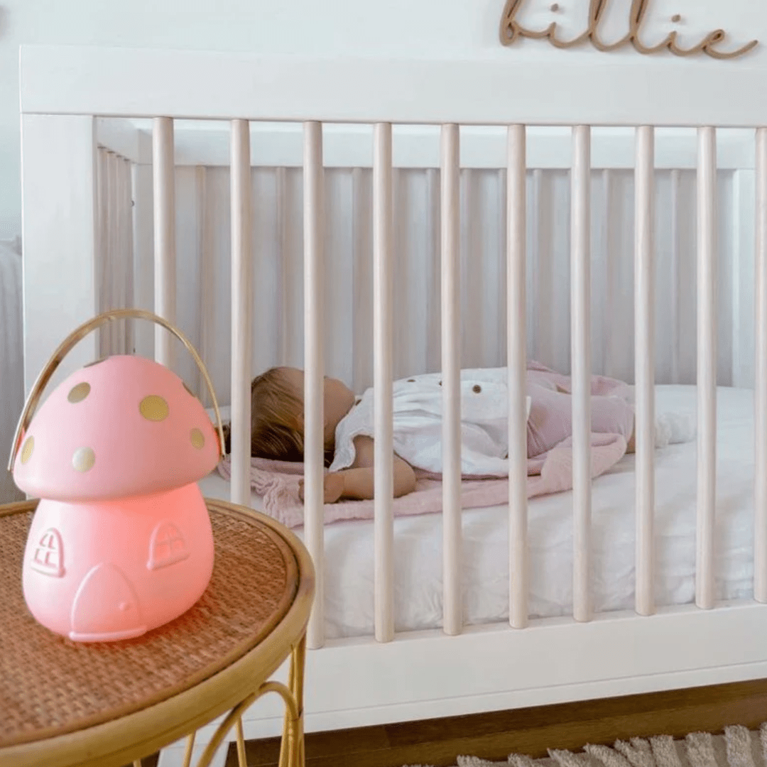 A toddler sleeping in a white crib beside a Little Belle Nightlights Mini Fairy House Carry Lantern on a wicker stool.