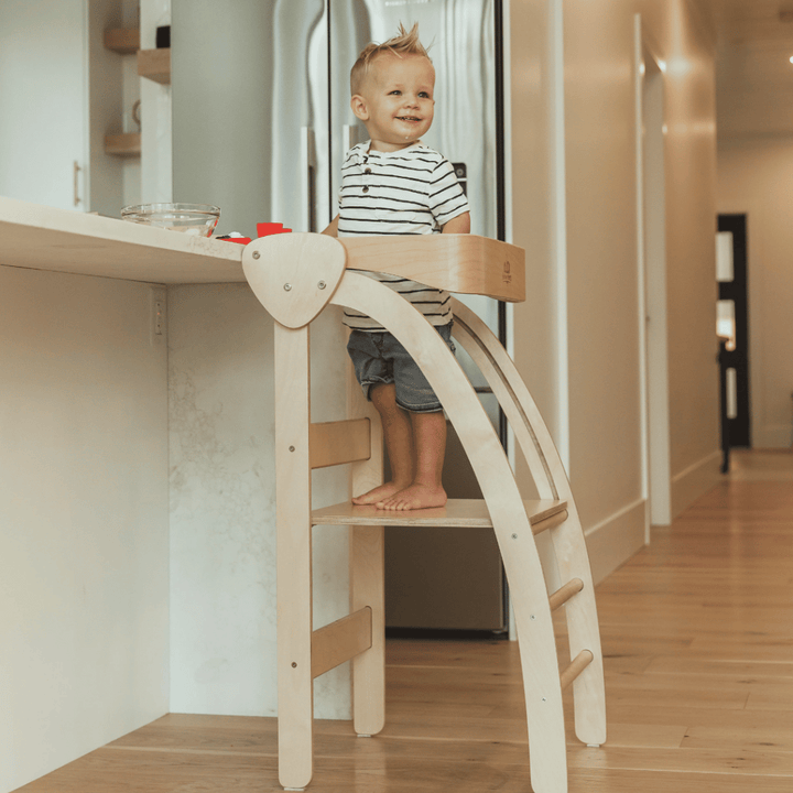 A child stands on the Kinderfeets Pikler Observation Tower, engaging in imaginative and functional play in a kitchen.