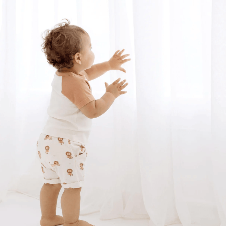 A baby, dressed in adorable Aster & Oak Organic Cotton Harem Shorts with an adjustable drawstring waist tie, is standing in front of a white curtain.
