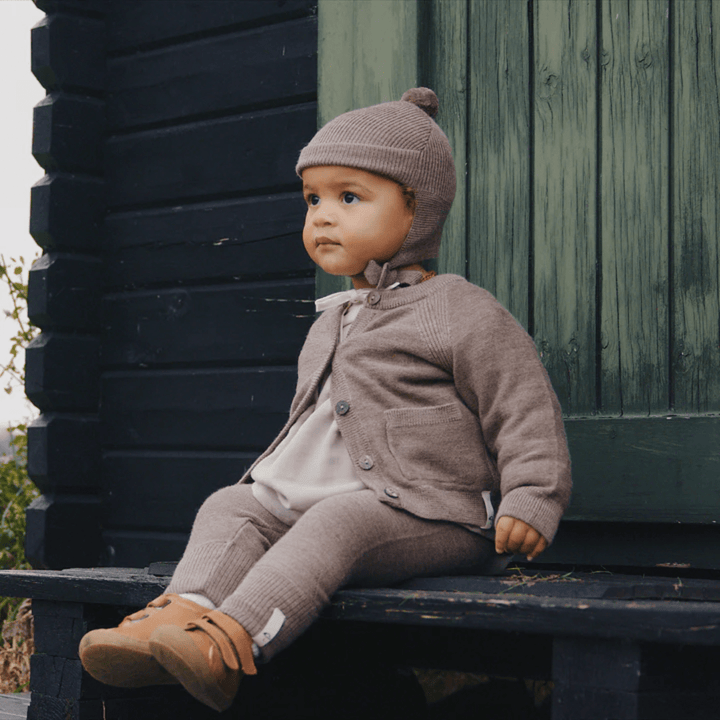 A baby sitting on a wooden bench wearing a grey sweater and Saga Copenhagen Merino Bonnet.
