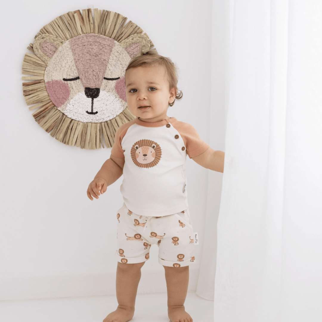 A baby wearing Aster & Oak organic cotton harem shorts, standing in front of a curtain with a lion on it.