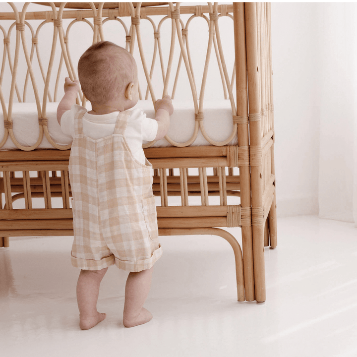 Back View Of Baby Boy Standing At Rattan Cot, Wearing A Cream Onesie & Taupe & Cream Gingham Muslin Short Overalls