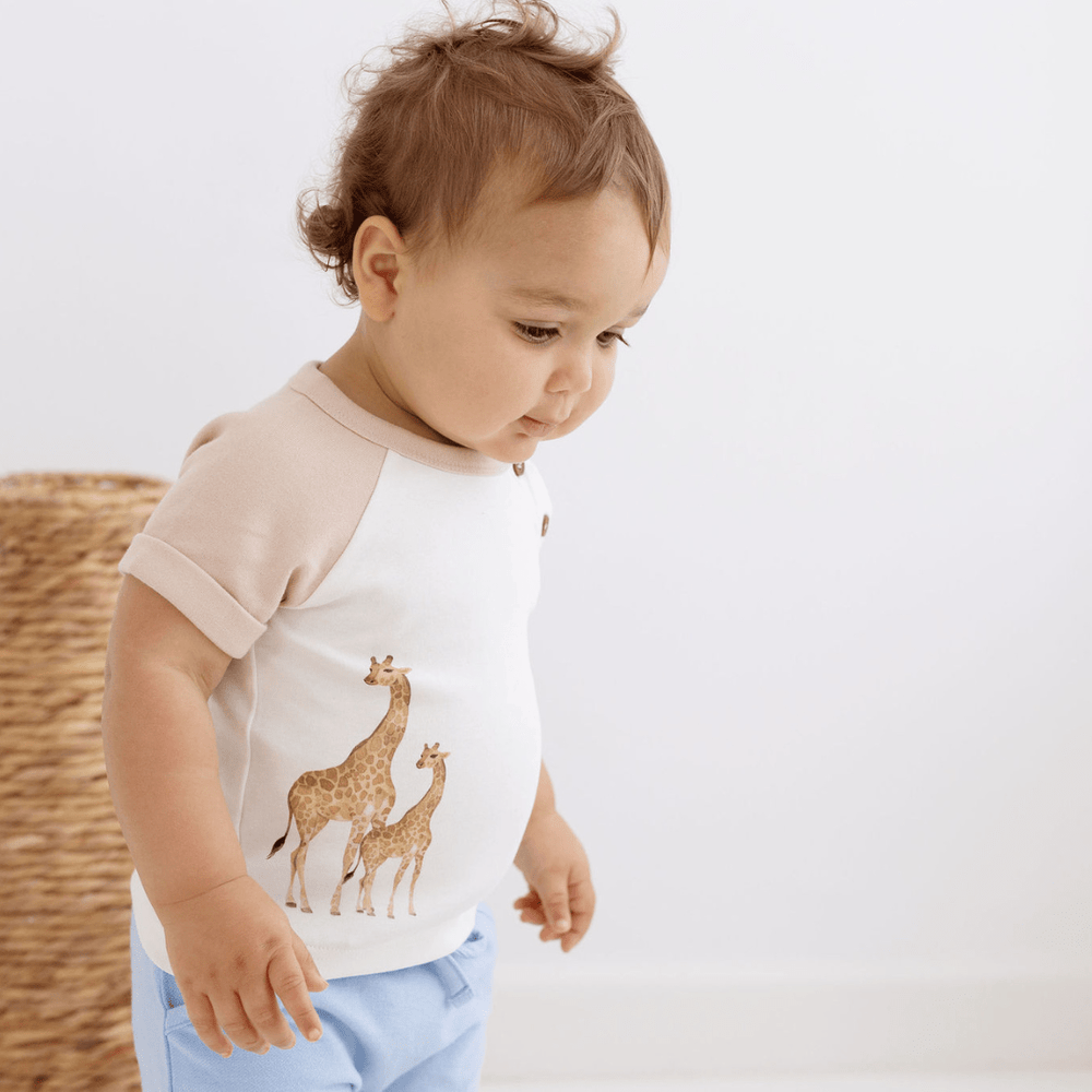 Brown-haired baby boy wearing a t-shirt with watercolour giraffe print on the right side, and contrasting taupe sleeves. He's wearing the t-shirt with blue shorts.