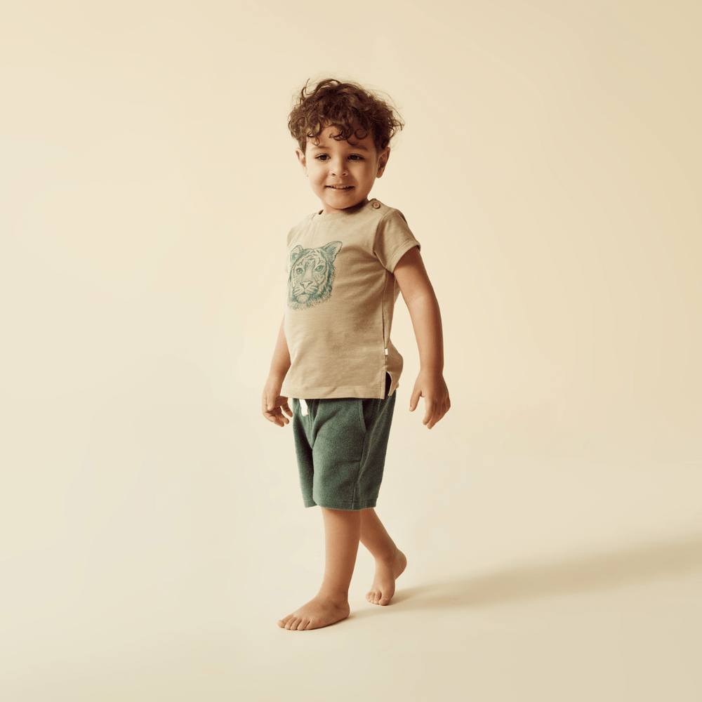 A young child with curly hair smiling and standing in a Wilson & Frenchy Leo the Lion Organic Kids Tee - LUCKY LAST - 4 YEARS ONLY, neutral-toned space.
