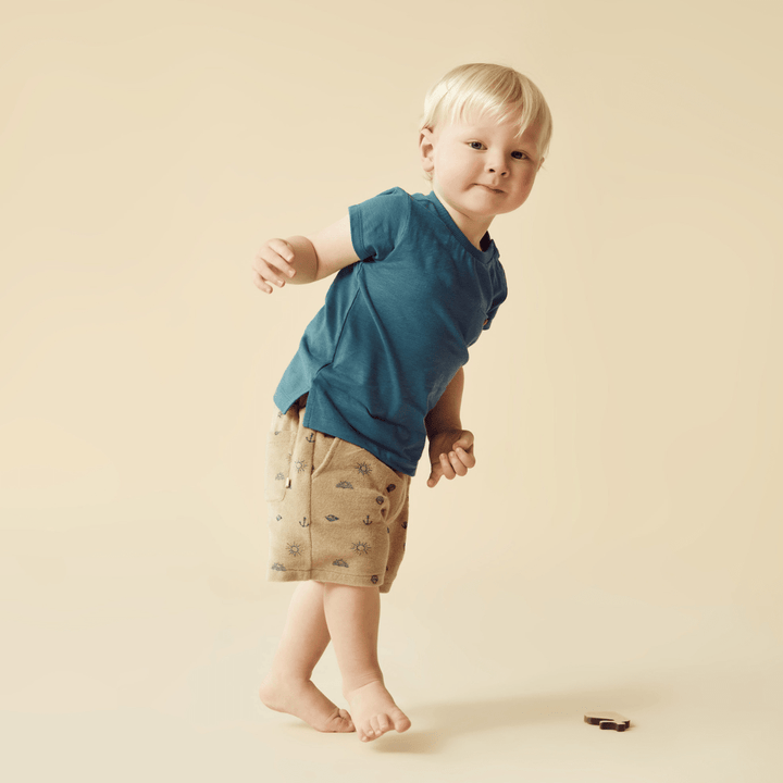 A young boy wearing a Wilson & Frenchy Organic Pocket Tee (Multiple Variants) and shorts on a beige background.