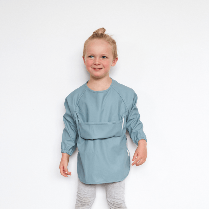 A little girl wearing a blue apron standing in front of a white wall, wearing Zazi Recycled Full-Sleeved Bibs (Multiple Variants).