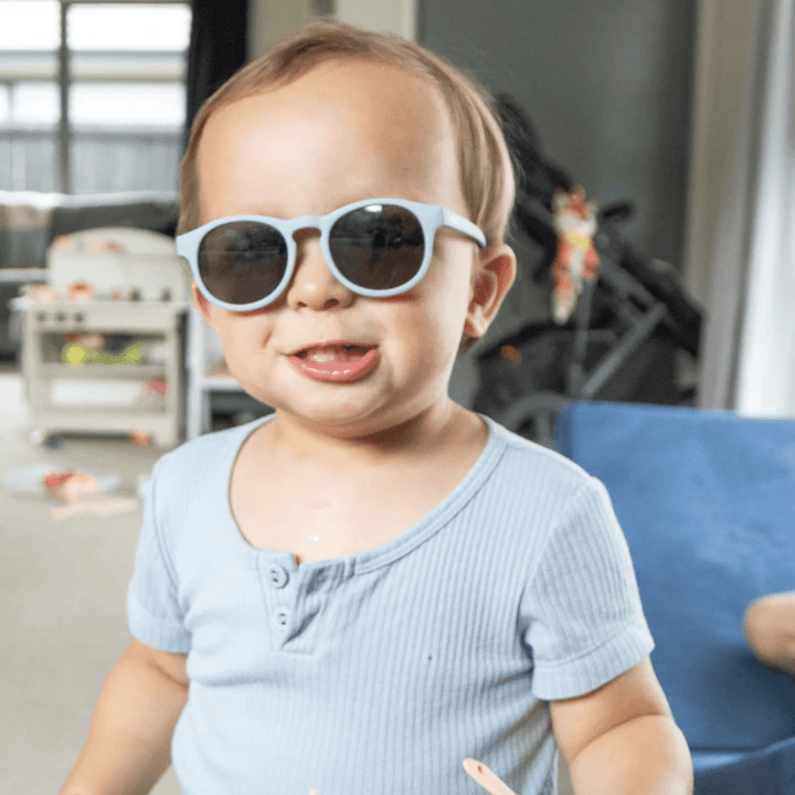 A baby wearing Zazi Shades Baby & Toddler Sunglasses by Zazi in a living room, with UV400 protection.